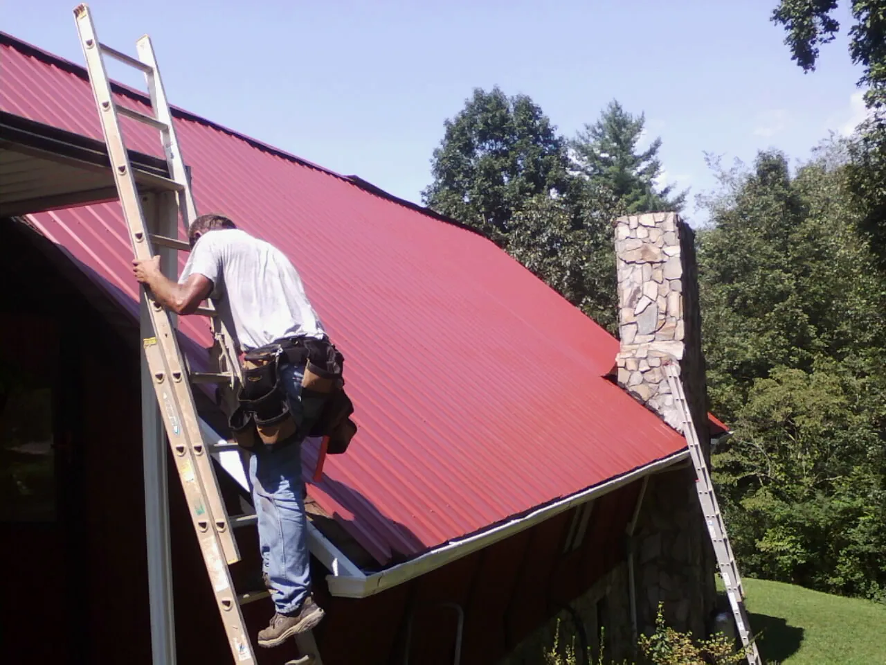 Roofer on a ladder beside a red metal roof and stone chimney — Soffit & Fascia Repair by Roofing Brothers in Ronald